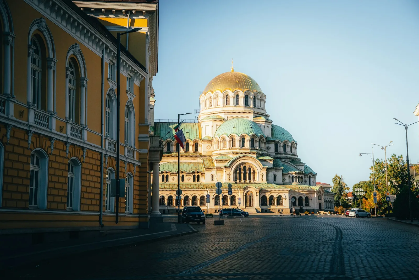The golden dome and green cupolas of Alexander Nevsky Cathedral in Sofia, viewed from a cobblestone street lined with historic buildings