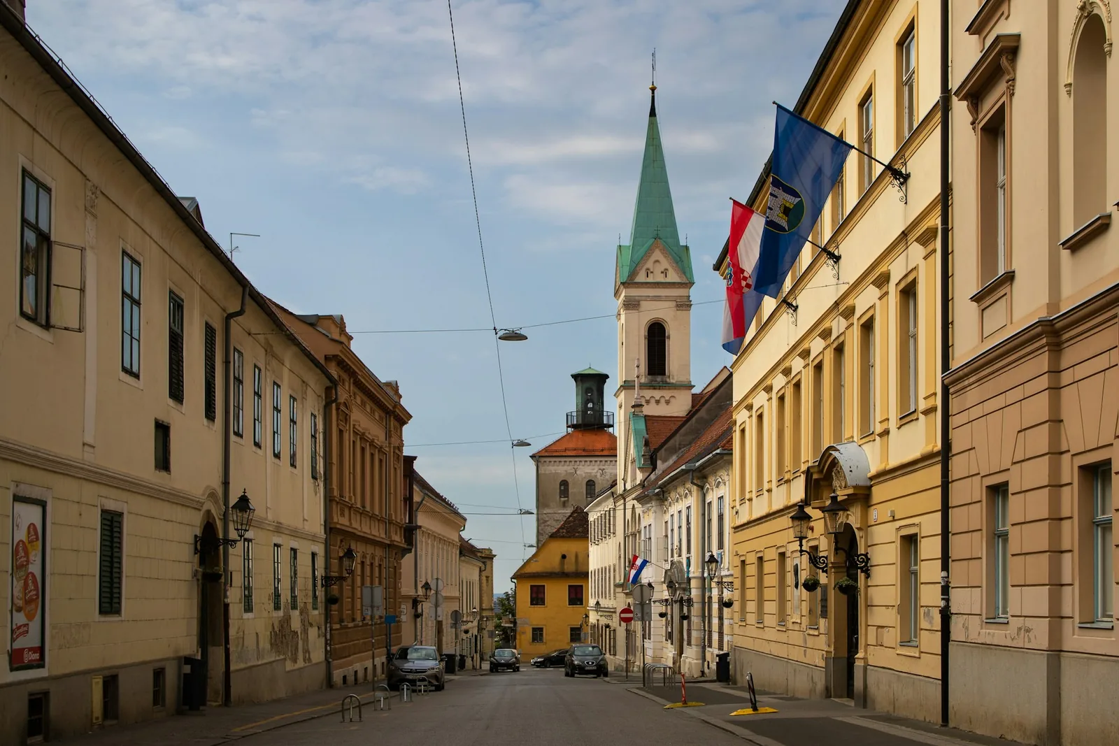 A Zagreb street lined with pastel townhouses leading to a church spire, the Croatian and city flags flying from a government building facade